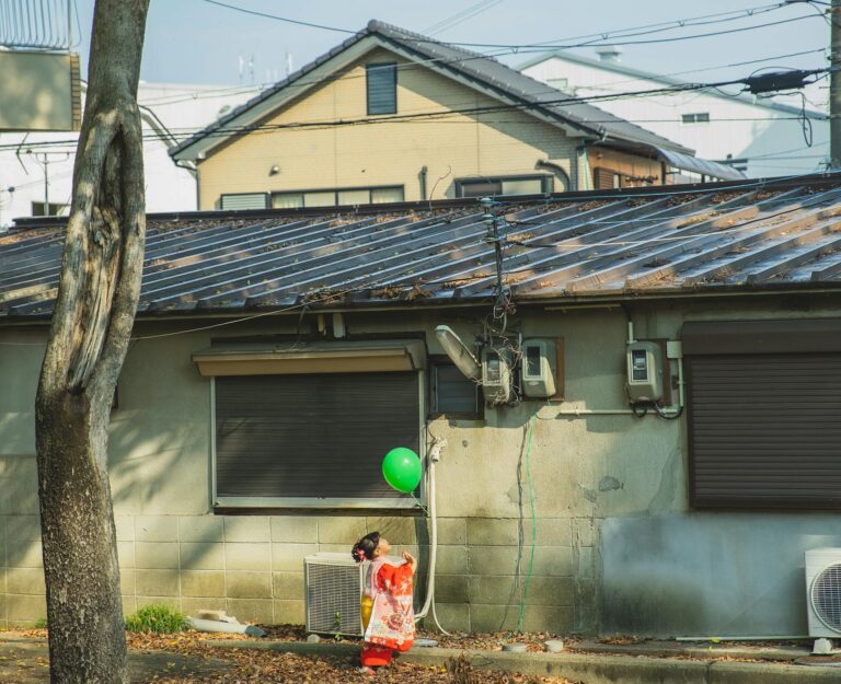 A child in a kimono holding a green balloon stands near a traditional Japanese house under a tree.